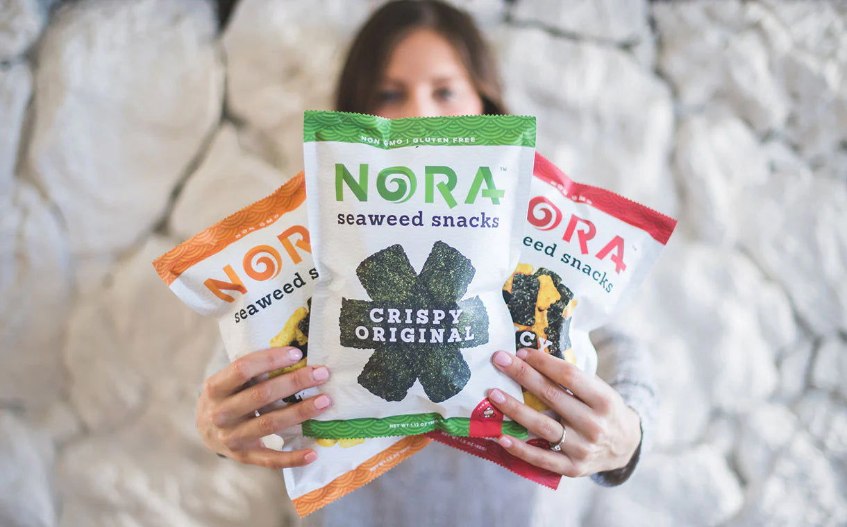 Young woman is holding up 3 different bags of Nora Seaweed Snacks