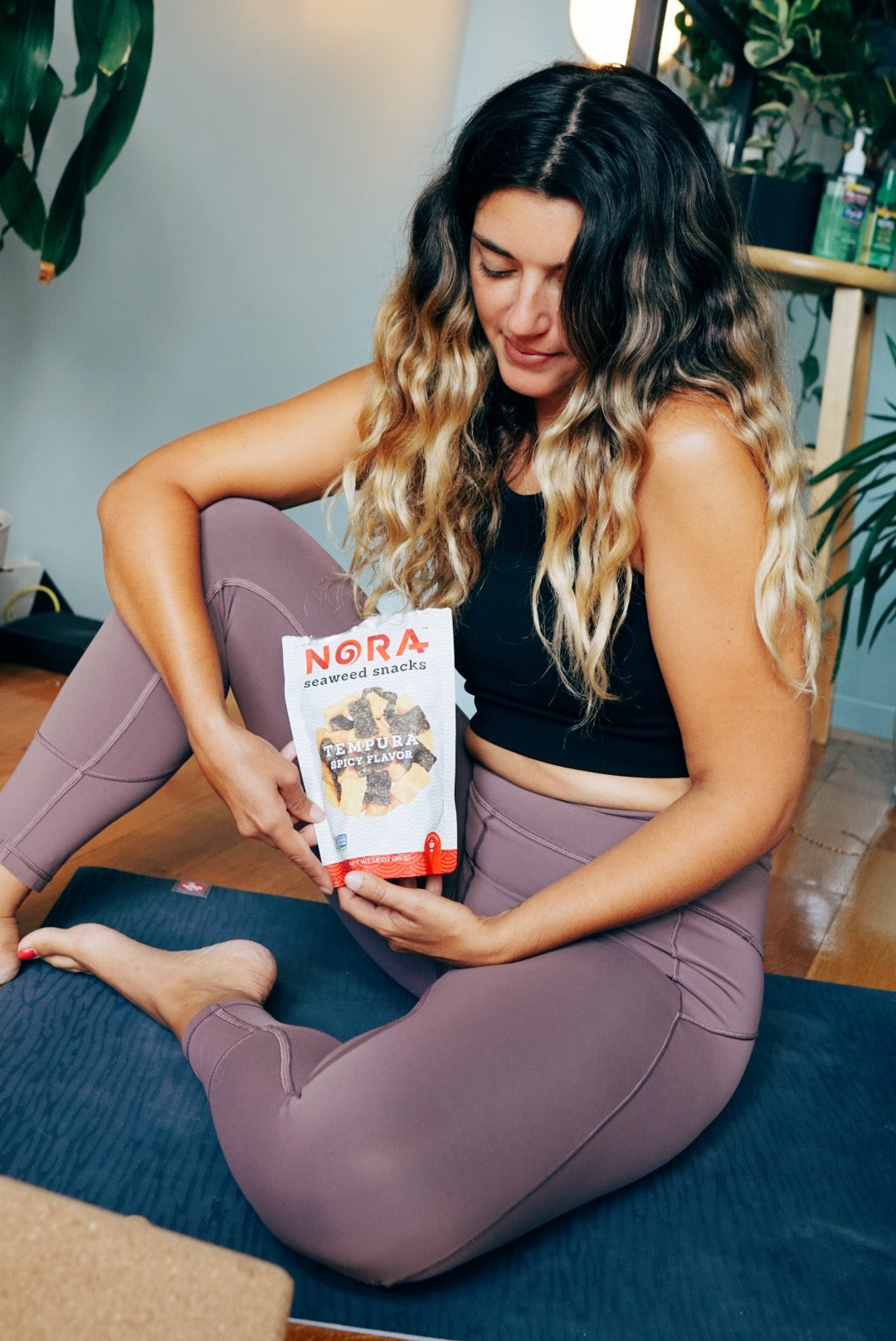 Young woman is holding Nora Seaweed Snacks bag while sitting on a floor.