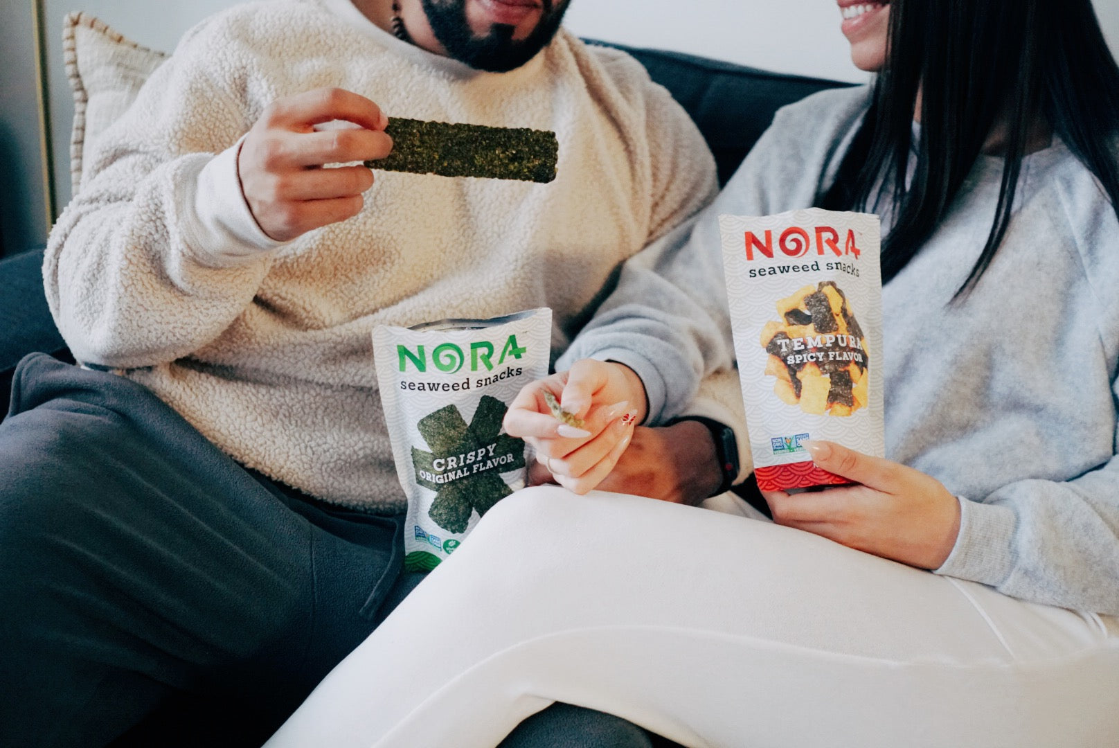 A couple is enjoying andd sharing Nora Seaweed Snacks on a couch